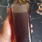A hand holds a tall bottle of dark honey with a black cap and honeycomb texture, matching Pure medium honey-Wild and organic.